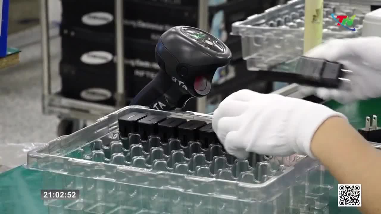 White-gloved hands pick up a black power adapter from a tray of identical items. A scanner hovers nearby, ready to record the product.