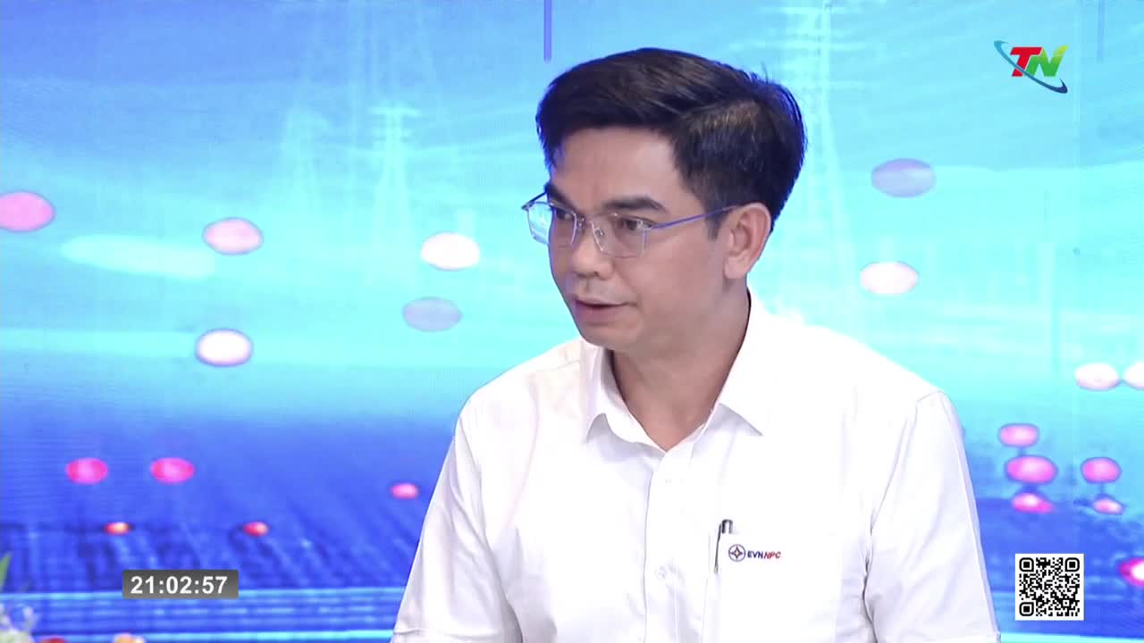 A man in a white shirt speaks on a television program, his glasses reflecting the bright studio lights. The background displays a digital pattern of soft, glowing orbs.