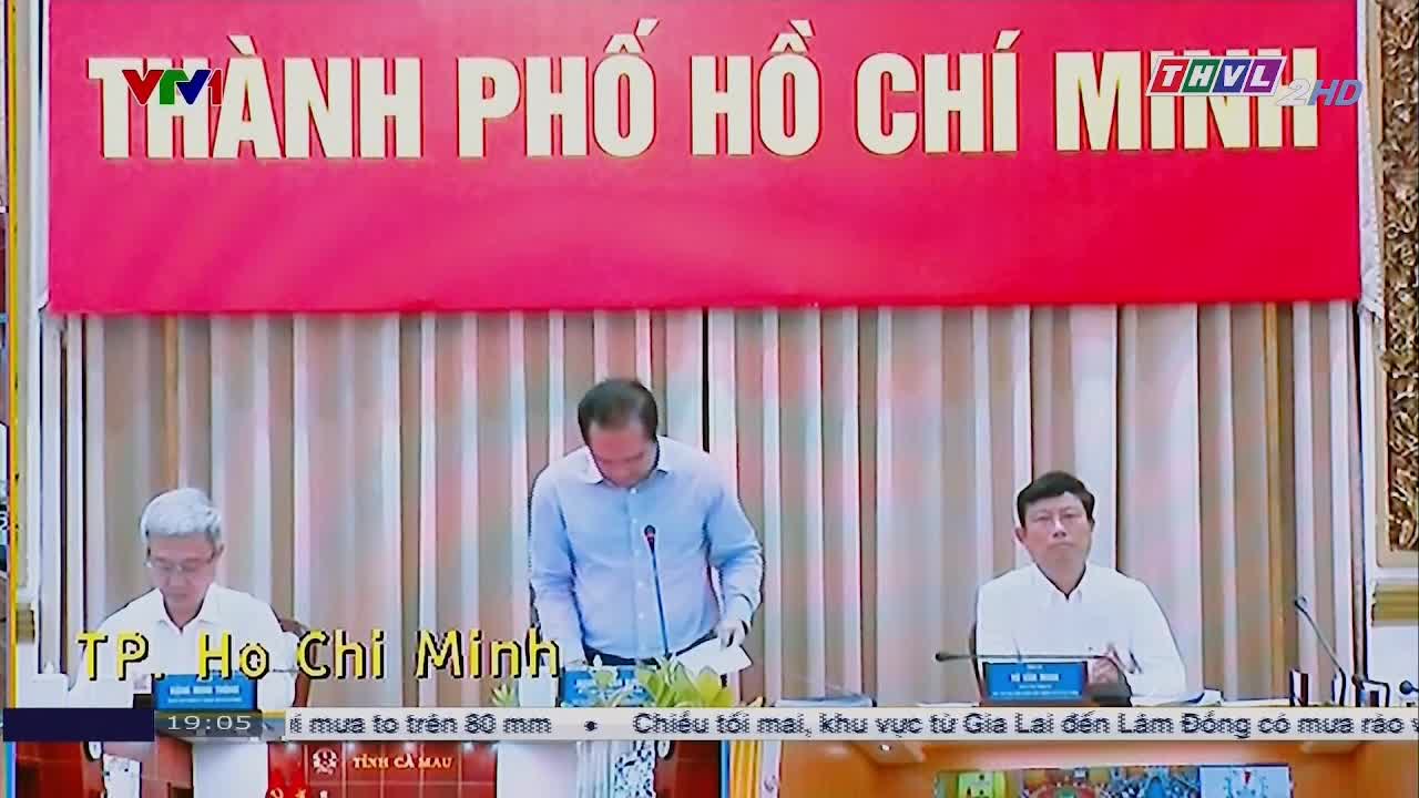 Three men sit at a long table, the central one leaning over papers. A red banner with Vietnamese text hangs above them, and the broadcast is on TV.
