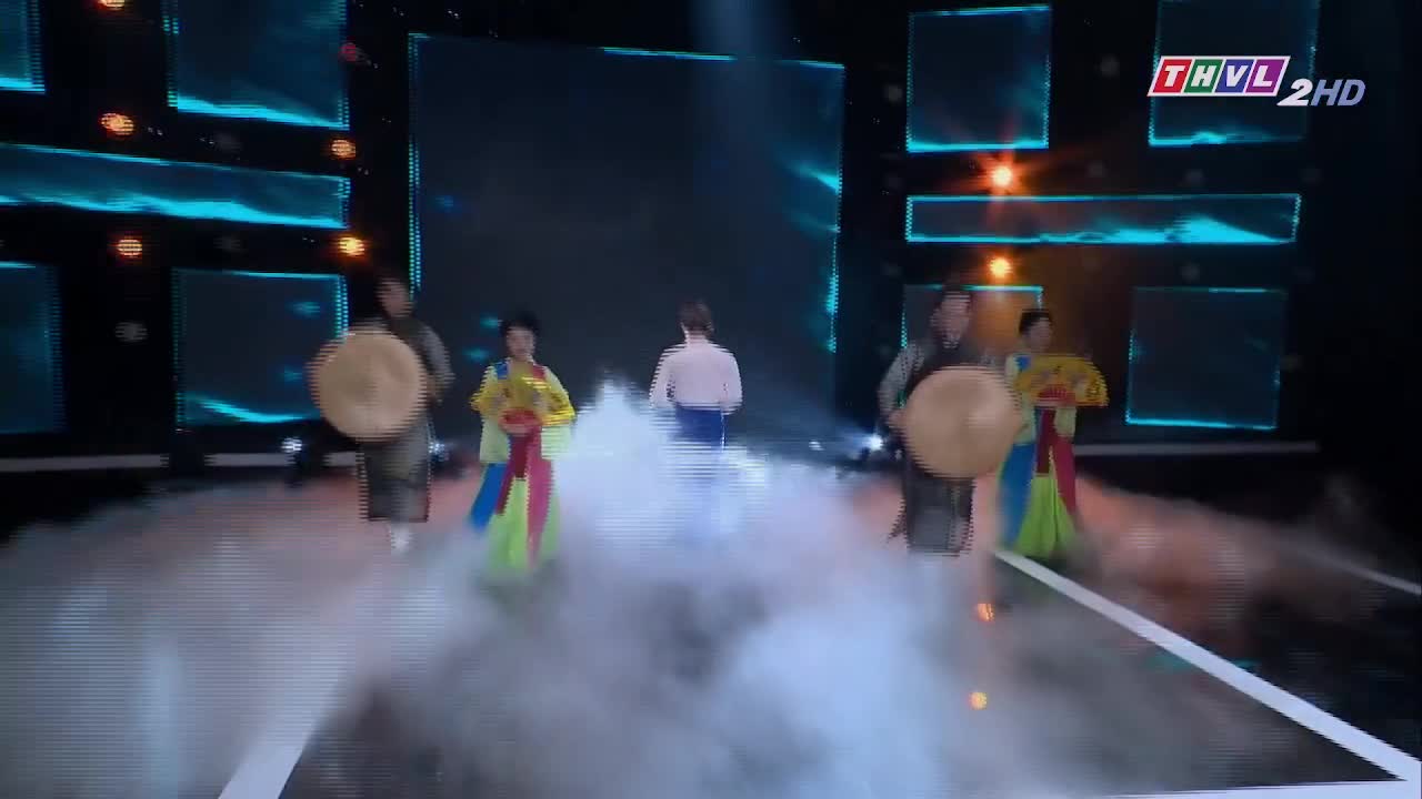 On the stage of Tra Vinh TV, performers in colorful traditional costumes and holding large, round drums move forward through a cloud of stage fog. A man in a white shirt walks in the center of the stage, facing away from the camera.
