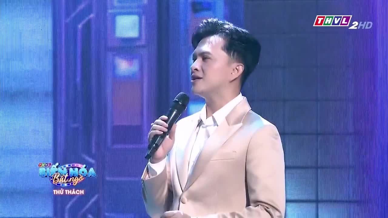 A man in a light-colored suit holds a microphone, singing on a stage lit with blue hues. The scene is likely a performance broadcast on Tra Vinh TV, based on the logos.
