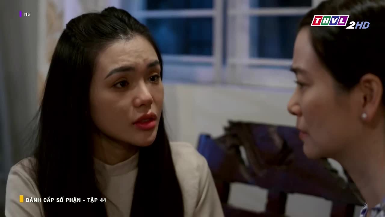 A woman with dark hair and red lipstick is speaking, her expression a mix of concern and sadness. Across from her, another woman listens intently, her face partially obscured.

