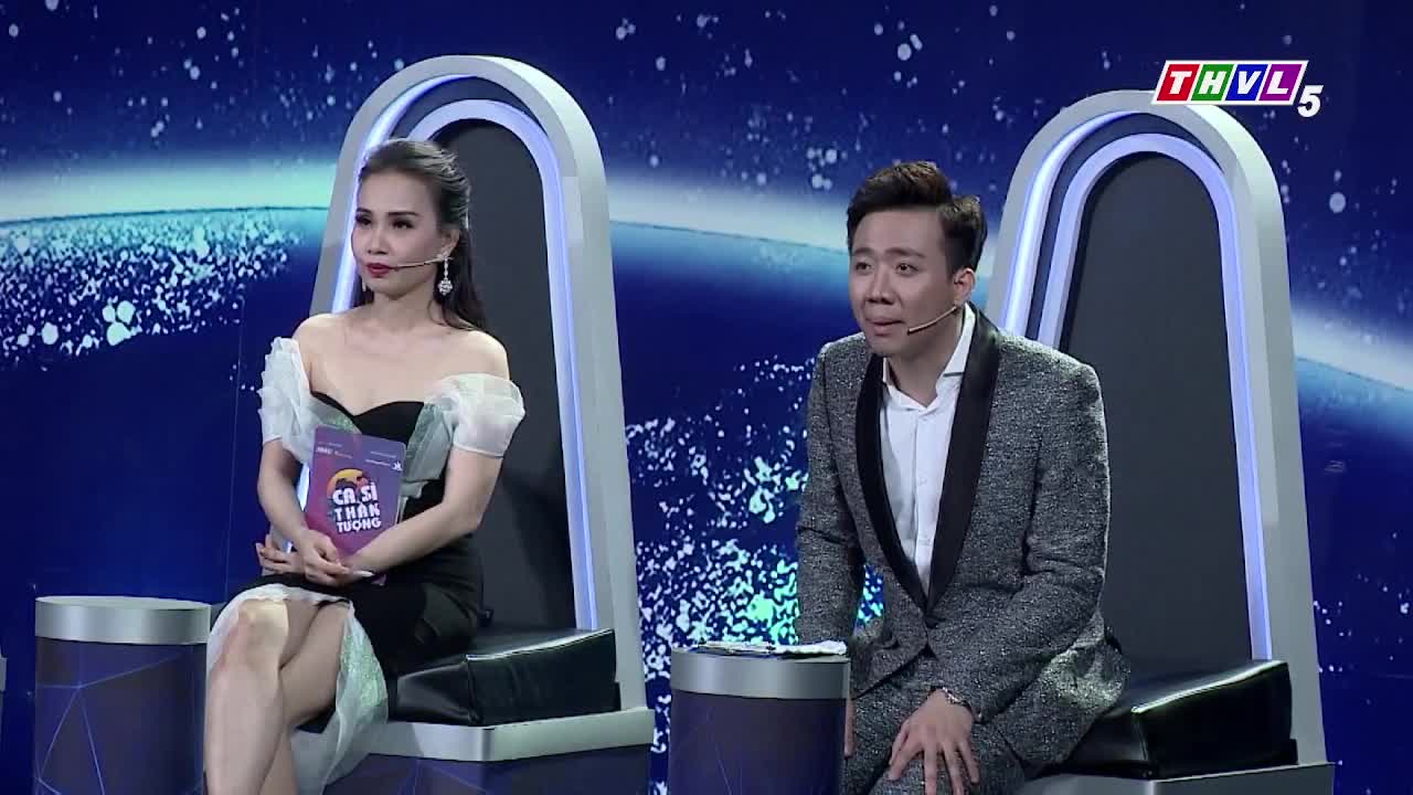 On a stage lit with blue, two people sit in tall, modern chairs, each with a microphone clipped to their lapels. The woman holds a book, while the man looks slightly surprised.
