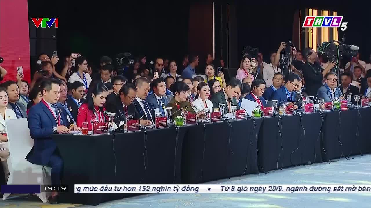 A panel of formally dressed people sits behind a long table, microphones in front of them. Behind them, a crowd of people holds up phones, likely recording the event for Tra Vinh TV.
