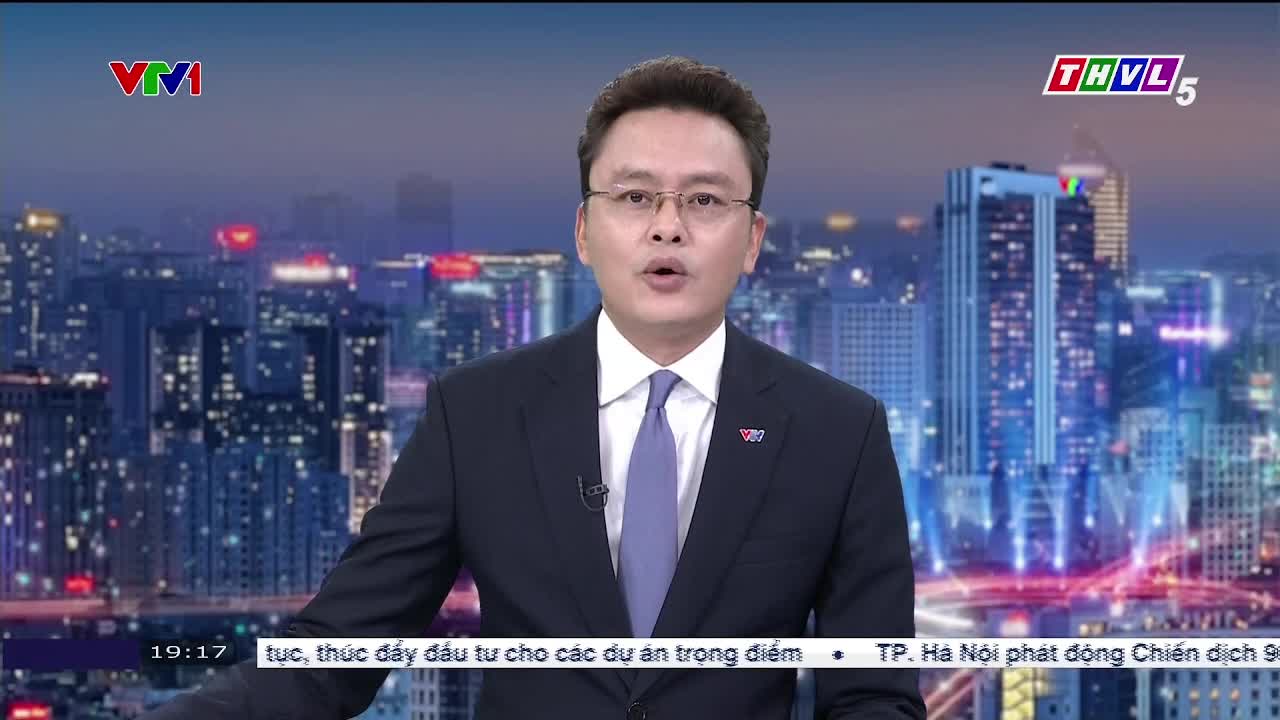 A man in a suit, wearing glasses, is speaking directly to the camera, his mouth open. The backdrop is a cityscape at night, with the logos of VTV1 and Tra Vinh TV visible.
