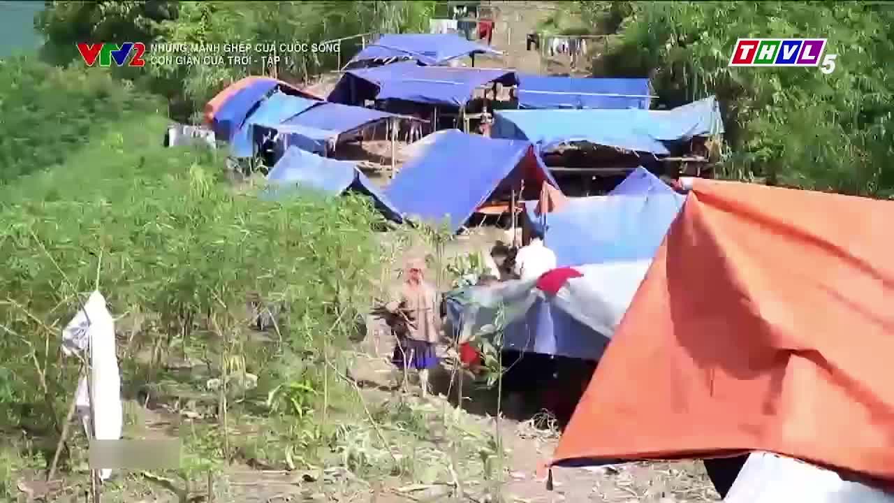 Tents, mostly blue, are pitched on a hillside, and people are milling about. A man in a long-sleeved shirt and shorts is walking towards the camera.
