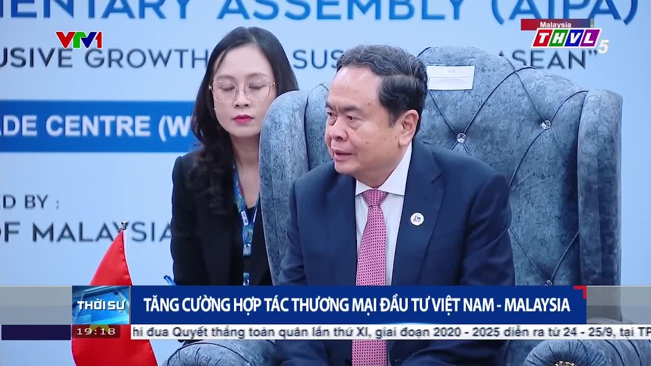 A man in a suit, with a pink tie, is speaking, his gaze directed to the side. A woman, wearing glasses, sits beside him, listening intently, as the Tra Vinh TV logo appears in the corner.
