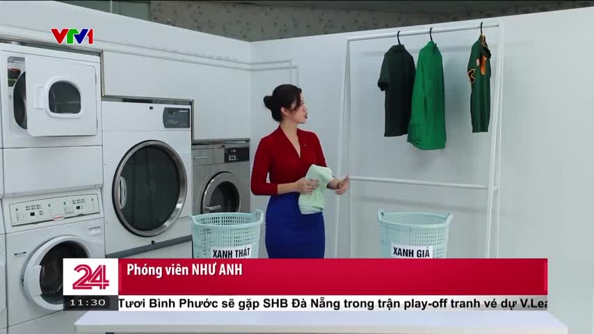 A woman in a red top and blue skirt gestures toward a rack of green shirts, holding a cloth. Behind her, several washing machines line the wall, and a VTV1 news banner is visible at the bottom.
