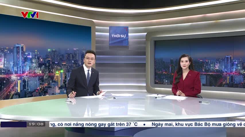 Two news anchors sit at a curved desk, facing the camera. The man in a suit gestures with a pen as he speaks, while the woman in a red blazer listens.

