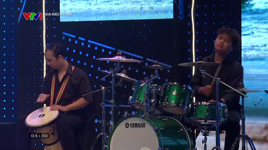 The man on the left slaps the conga drum with his hands, while the other drummer hits the cymbals and snare. The stage is lit with blue lights, and the Yamaha drum kit has a shiny green finish.
