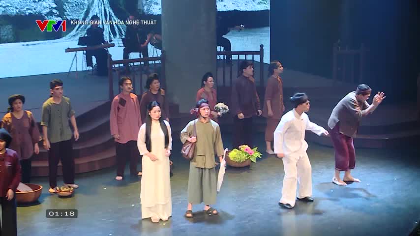 A group of actors in traditional Vietnamese clothing stand on a stage, some facing forward and others interacting with each other. One man in white gestures emphatically, while another in brown raises his hands in a pleading manner.
