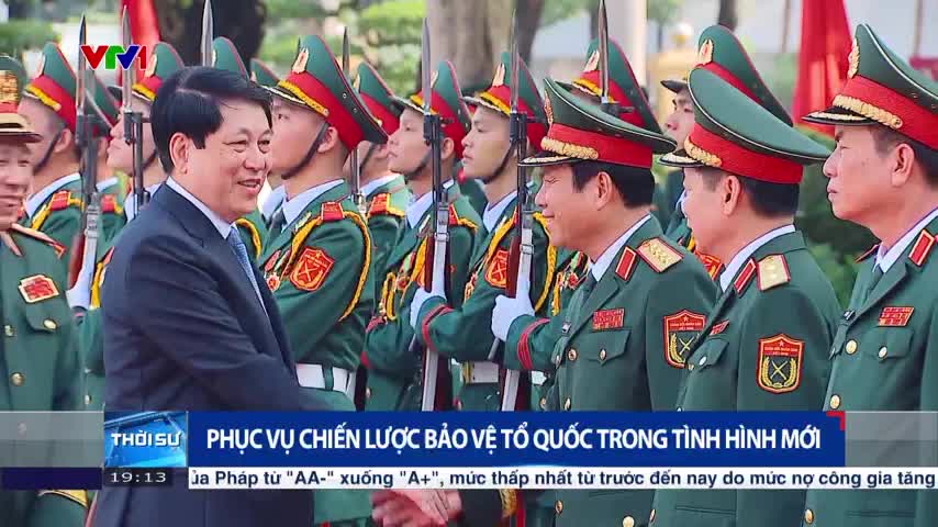 A man in a suit shakes hands with a man in a military uniform, both smiling. Behind them, soldiers in green uniforms and red-trimmed hats stand at attention, rifles held at their sides.
