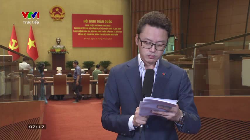 A man in a suit and glasses, holding a microphone, reads from a stack of papers. Behind him, in a large hall, people mill about near a stage with flags and a bust.
