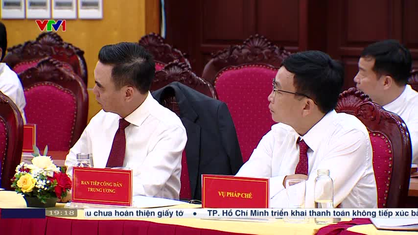 Men in white shirts and red ties sit at a long table, listening intently.  A VTV1 logo is visible in the corner, and text scrolls along the bottom of the screen.

