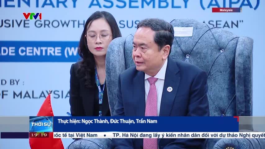 A man in a dark suit and pink tie speaks, looking slightly to his right. A woman in glasses sits beside him, listening intently, as the VTV1 news program rolls on.
