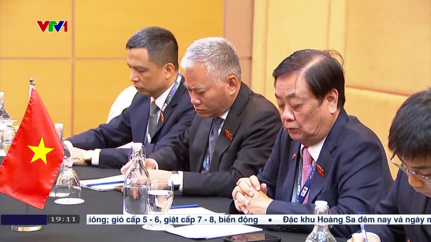 Men in dark suits sit at a table, focused on something out of view. A Vietnamese flag stands at the edge, and the VTV1 logo is visible on the screen.
