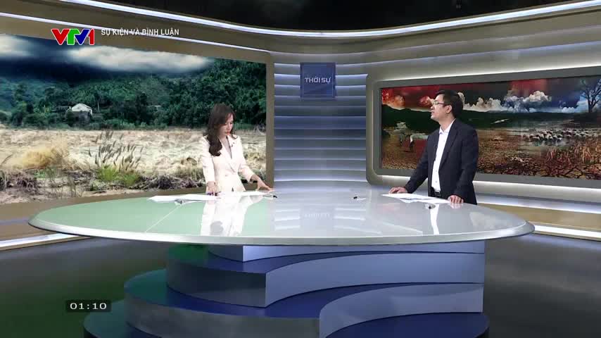 A woman in a white suit stands at a large, curved desk, gesturing towards a screen showing a flooded landscape. Across from her, a man in a dark suit leans forward, looking at the same screen, likely discussing the news on VTV1.
