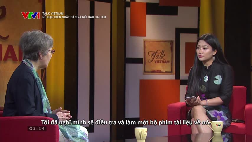 On a VTV1 set, a woman with long black hair sits holding a small device, listening intently. Across from her, an older woman with glasses and gray hair speaks, her hands gesturing slightly as she talks.
