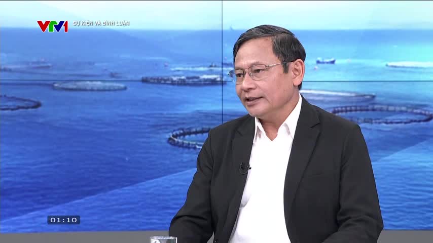A man in a black suit and glasses is speaking on VTV1, the backdrop showing a body of water with circular structures. The clock in the corner reads 01:10.
