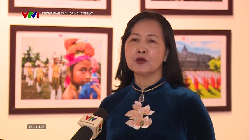 A woman in a blue áo dài speaks directly into a VTV1 microphone, framed by pictures on the wall behind her. The clock on the screen reads 01:12.
