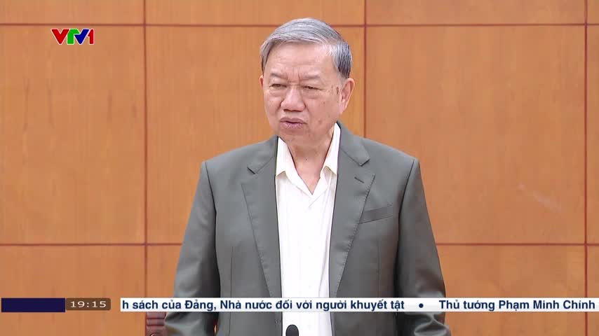 A man in a gray jacket is speaking, his expression serious. The VTV1 logo is visible in the corner, and a news ticker scrolls along the bottom of the screen.
