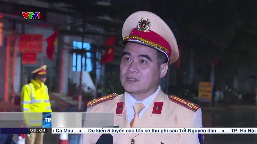 A traffic police officer in a light-colored uniform speaks directly to the camera, his red and gold insignia sharp against the dim evening light. Behind him, another officer in a bright yellow vest stands near a building adorned with Vietnamese flags.